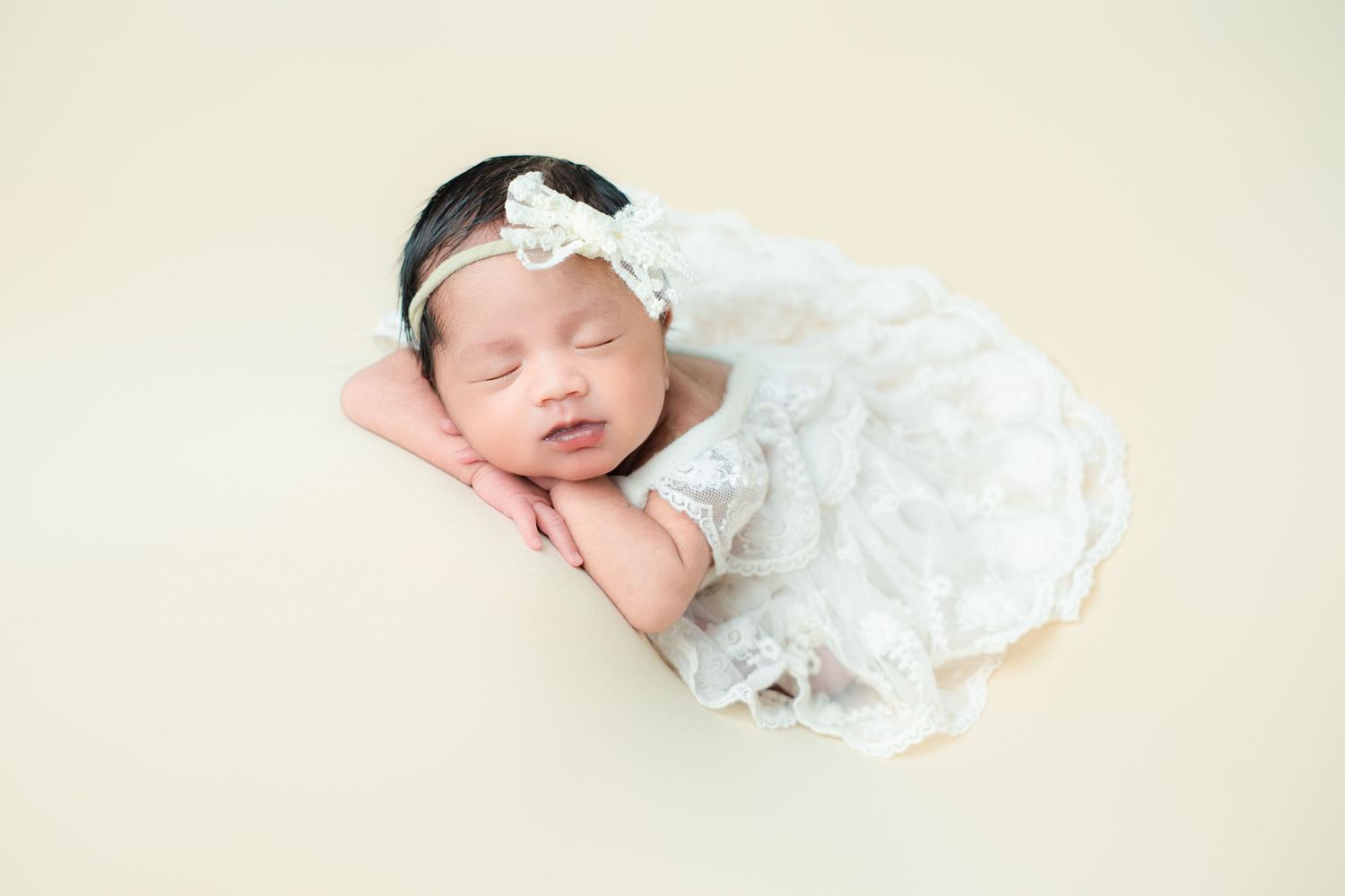 Peaceful sleeping newborn in white lace dress and headband on cream backdrop.