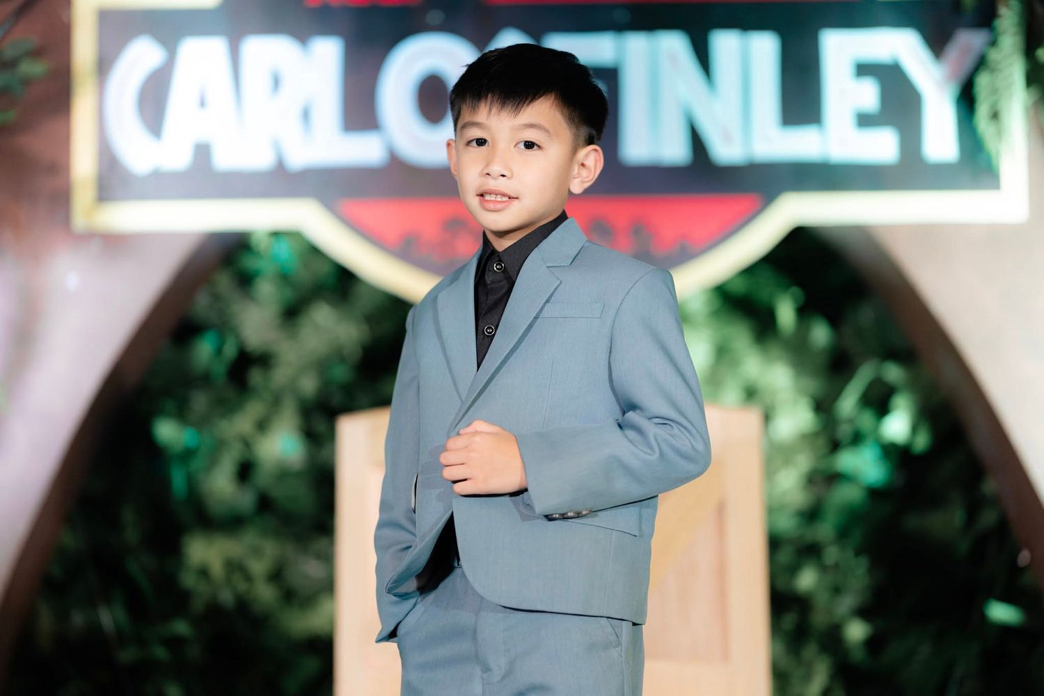 Young boy in gray suit poses confidently at Carlo Gatinley fashion event backdrop.