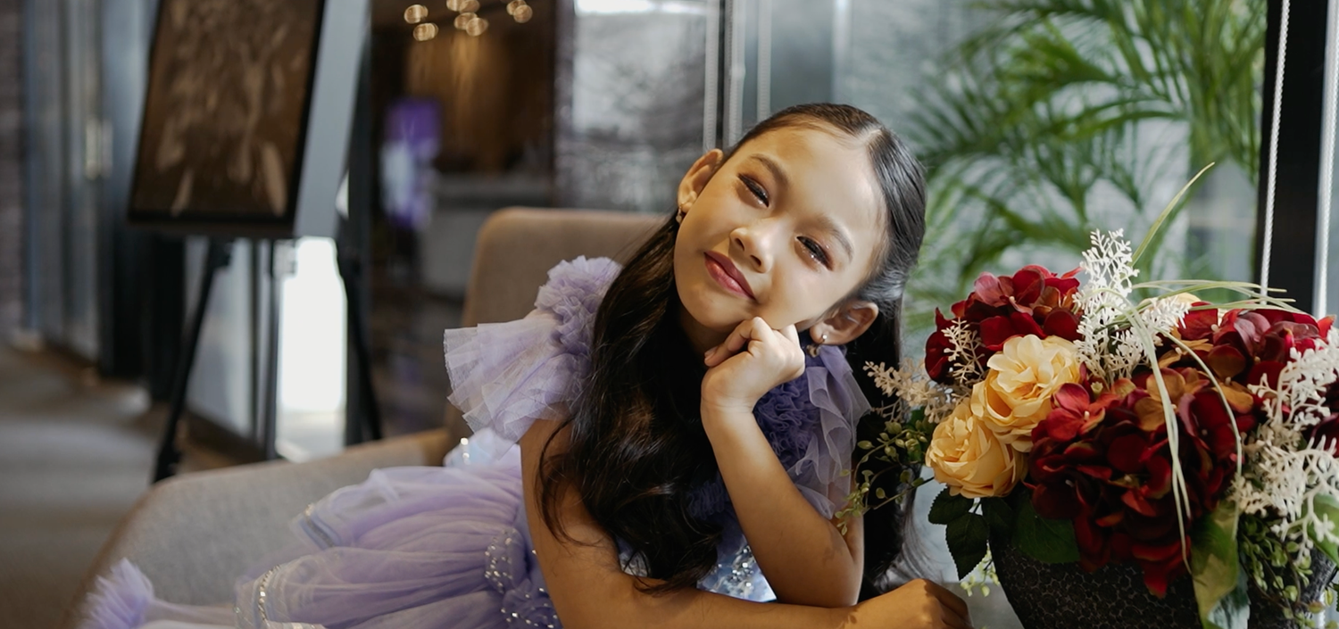 Young girl in purple tulle dress poses elegantly beside colorful floral arrangement in a softly lit room.