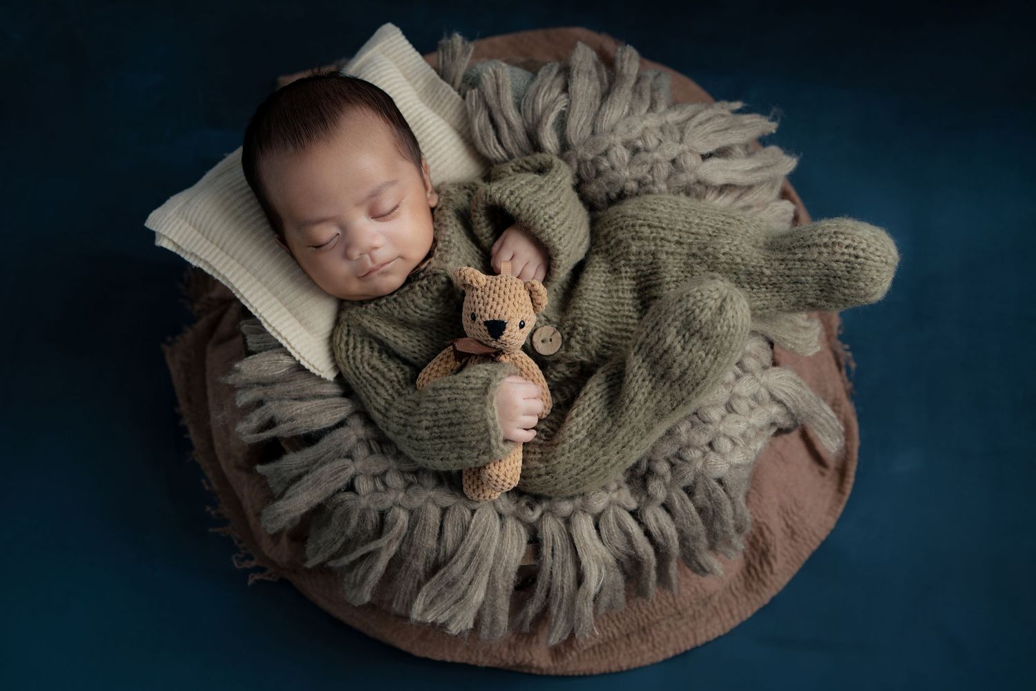 A sleeping baby curled up in a cozy round nest of soft gray fabric in a dark, moody setting.