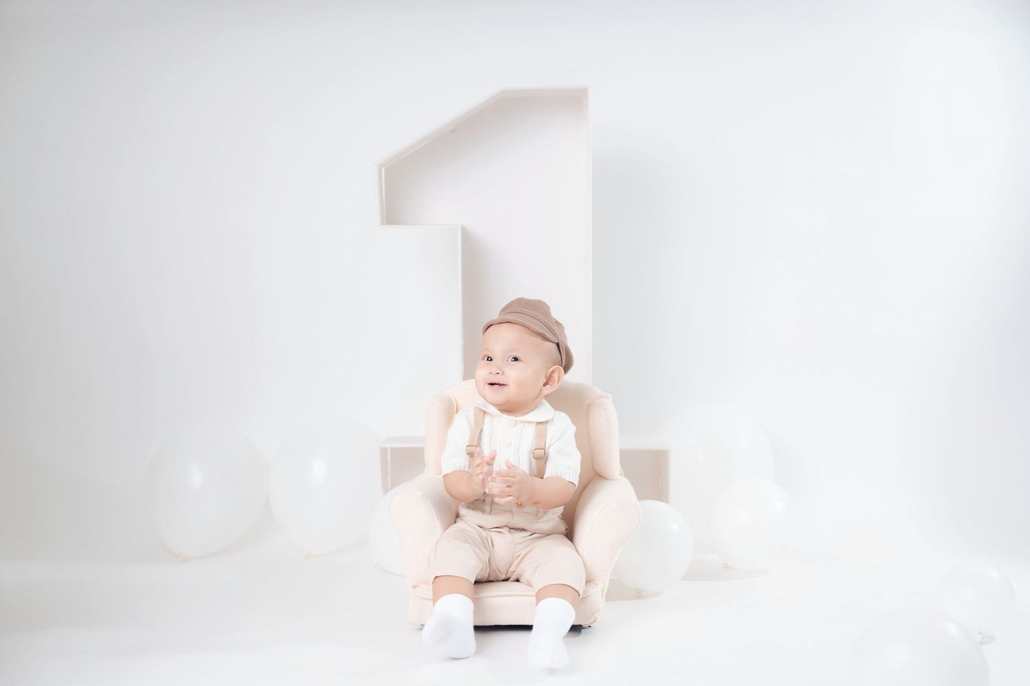 A baby in a white onesie sits on a white chair against a bright white minimalist background.