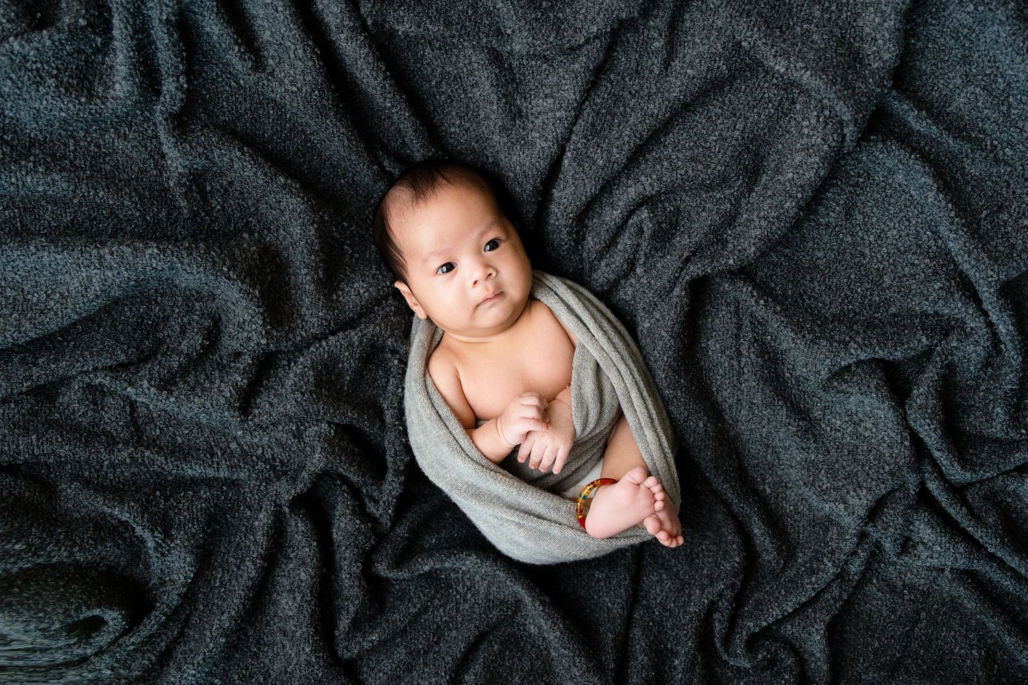 Swaddled baby laying on dark textured fabric backdrop during studio photoshoot.