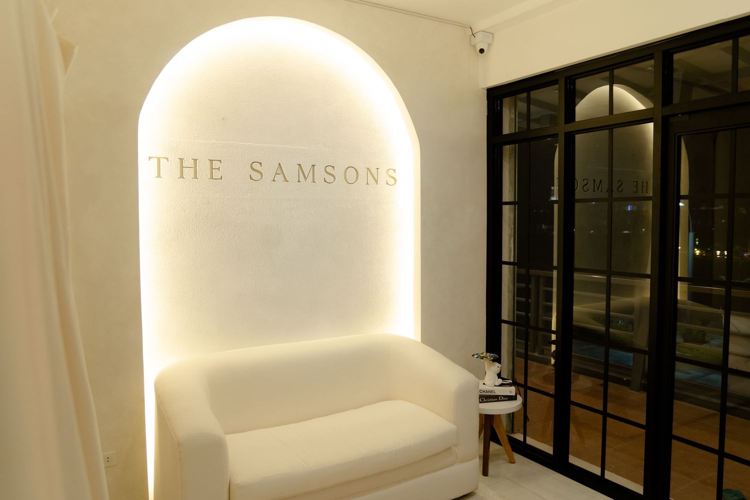 White arched alcove with backlit text reading 'THE SAMSONS' above a white loveseat against a dark backdrop.
