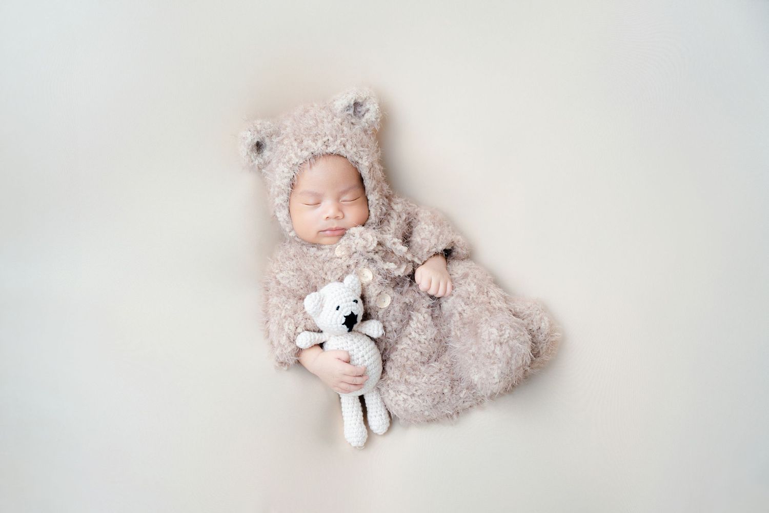 Newborn in fuzzy bear outfit with plush toy on cream background.