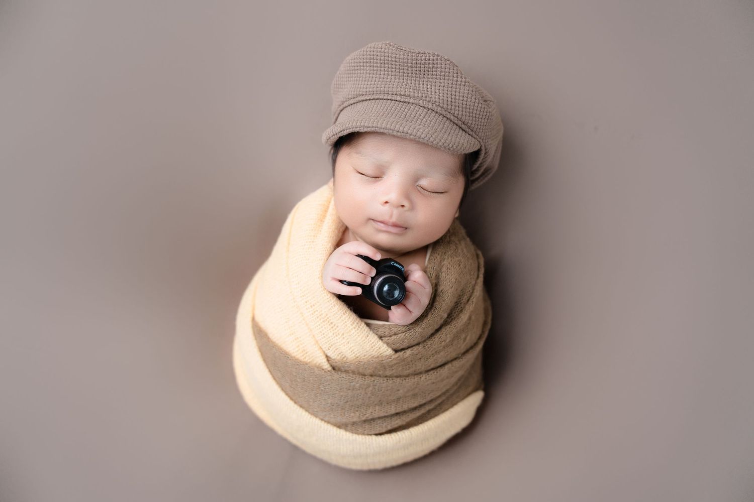 Sleeping baby wrapped in beige fabric with matching newsboy cap.