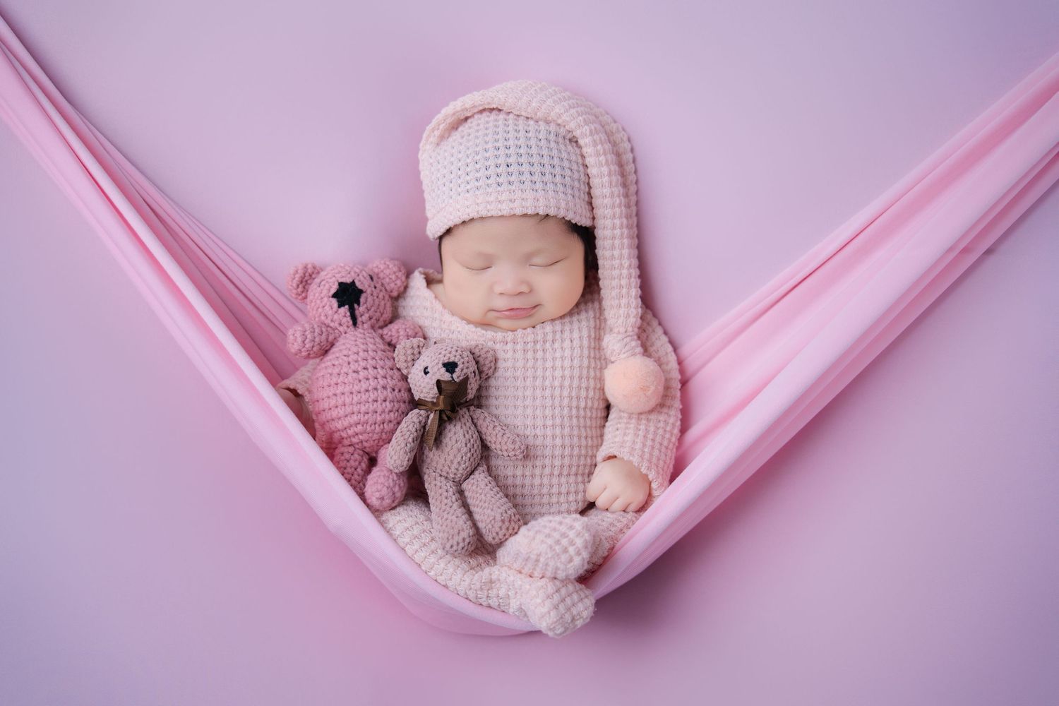 Sleeping newborn in knit outfit with teddy bear in pink fabric hammock.