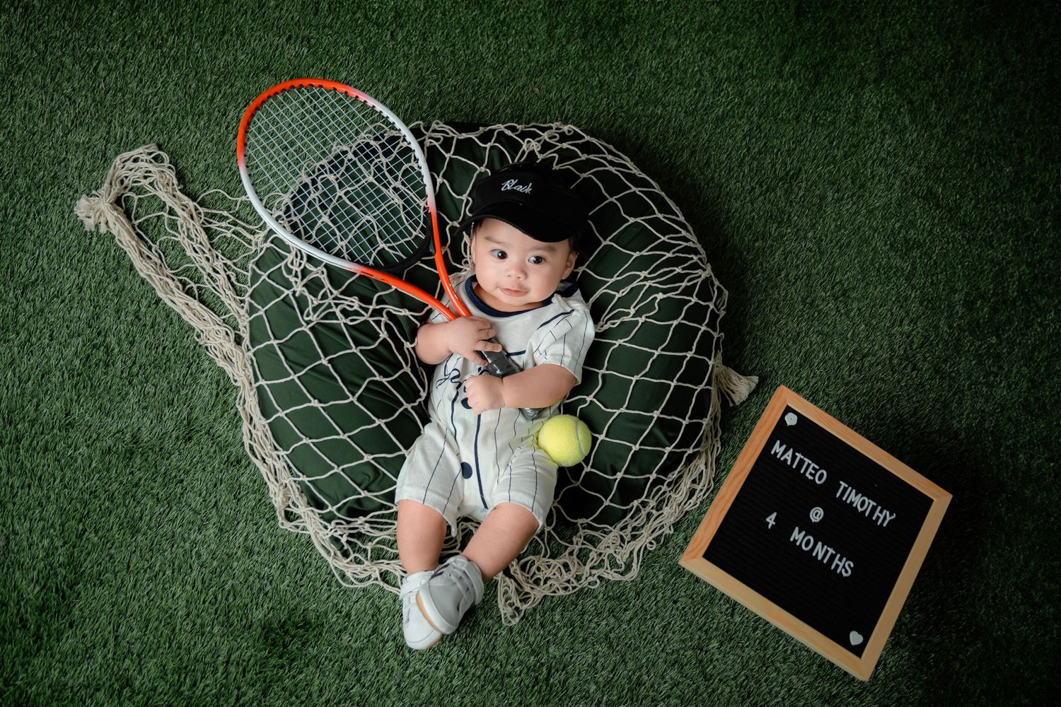 A tennis-themed photo setup on green turf with a tennis racket, ball, and net backdrop.