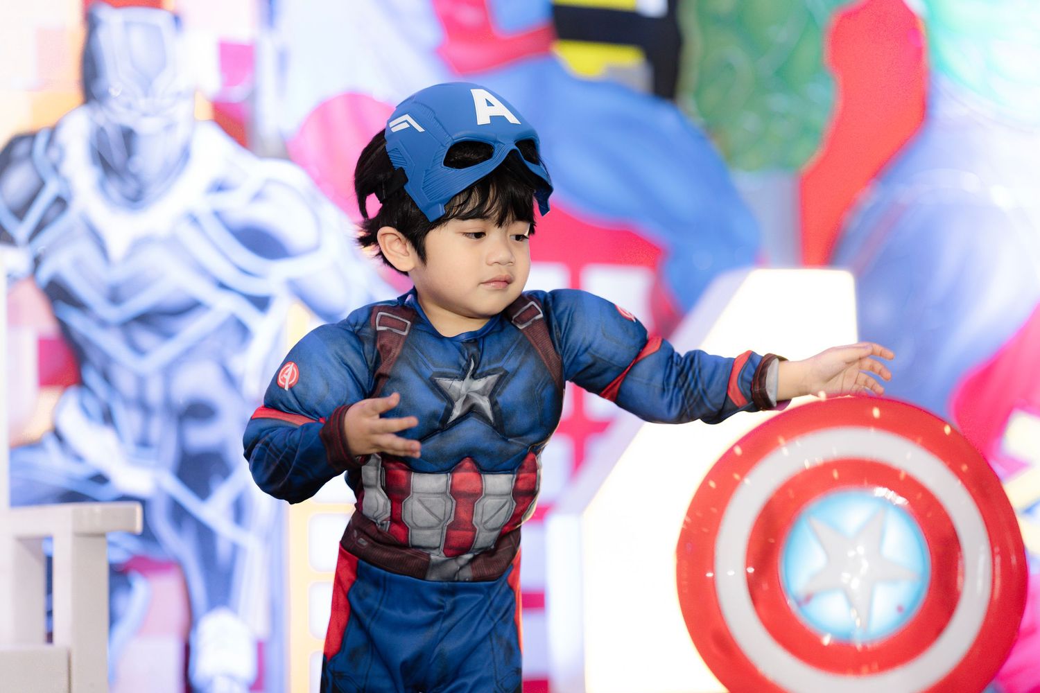 Young boy dressed in Captain America costume with shield and helmet at Marvel-themed birthday party.