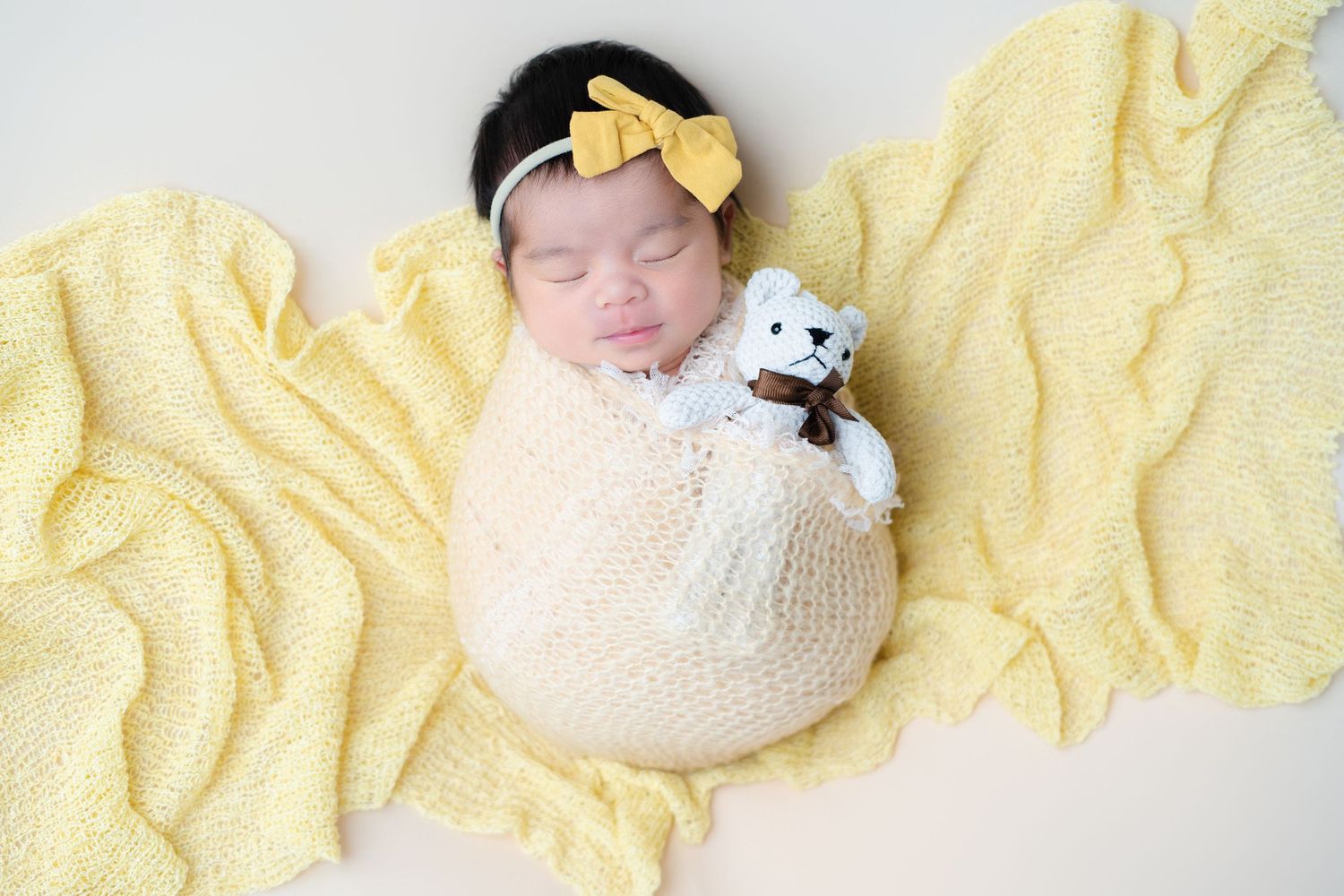 Sleeping newborn with yellow bow wrapped in cream knit on yellow blanket with small plush toy.