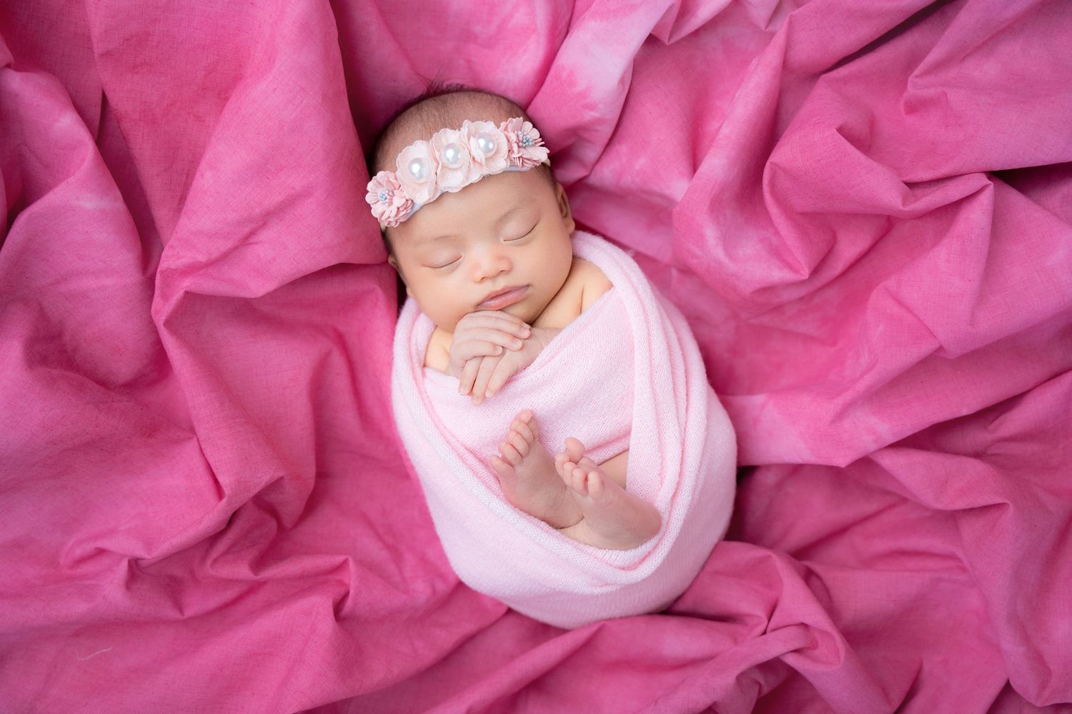 Sleeping newborn wrapped in white swaddle on pink textured fabric with lace headband.