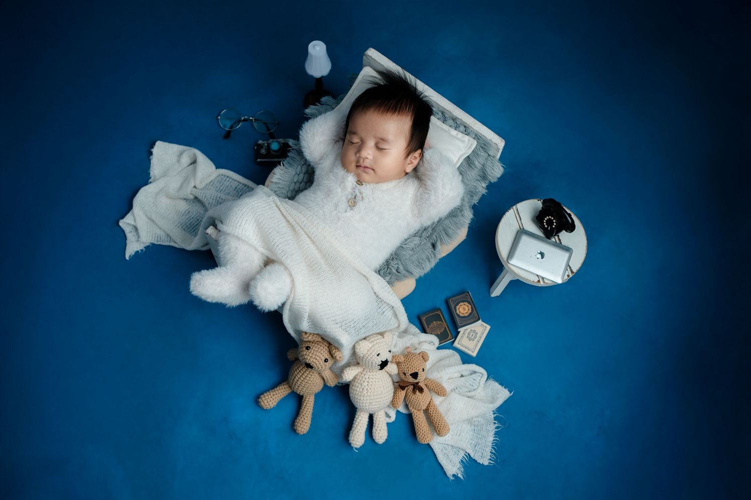 Sleeping baby in white clothing surrounded by stuffed animal toys on blue background.