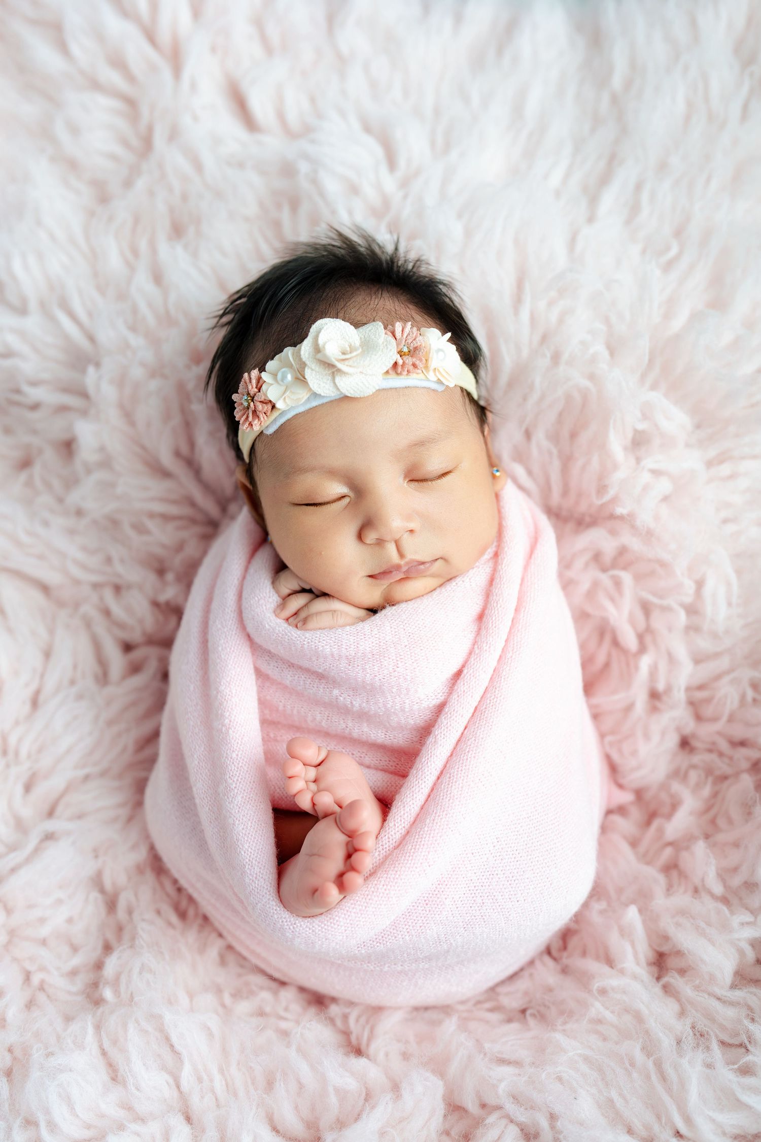 Sleeping newborn wrapped in soft pink blanket wearing floral headband on fluffy pink fur background.