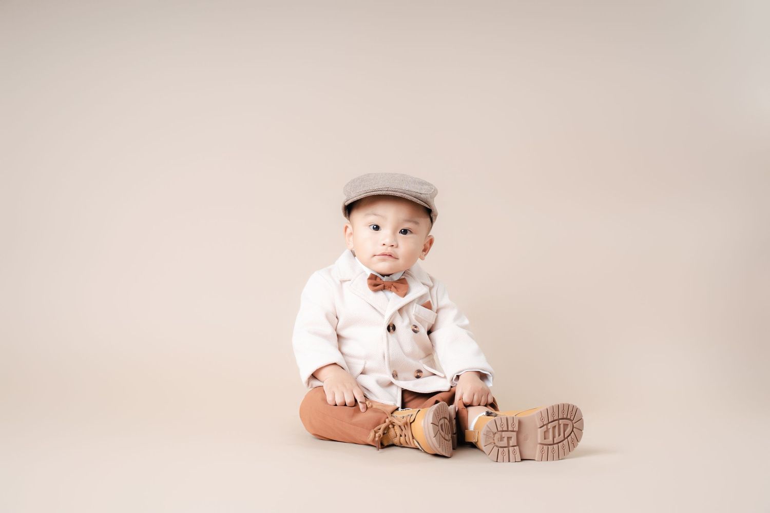 Baby wearing beige newsboy cap, white sweater and brown pants sits on cream backdrop with knit booties.
