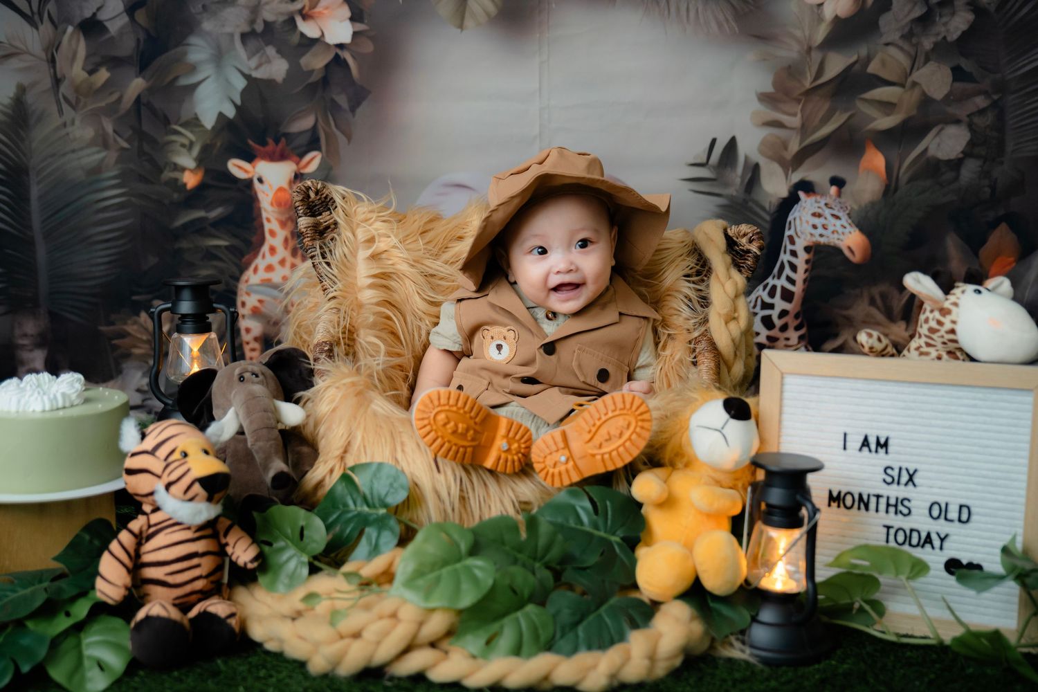 Safari themed baby photography setup with plush animals, lantern and greenery in a dreamy woodland setting.
