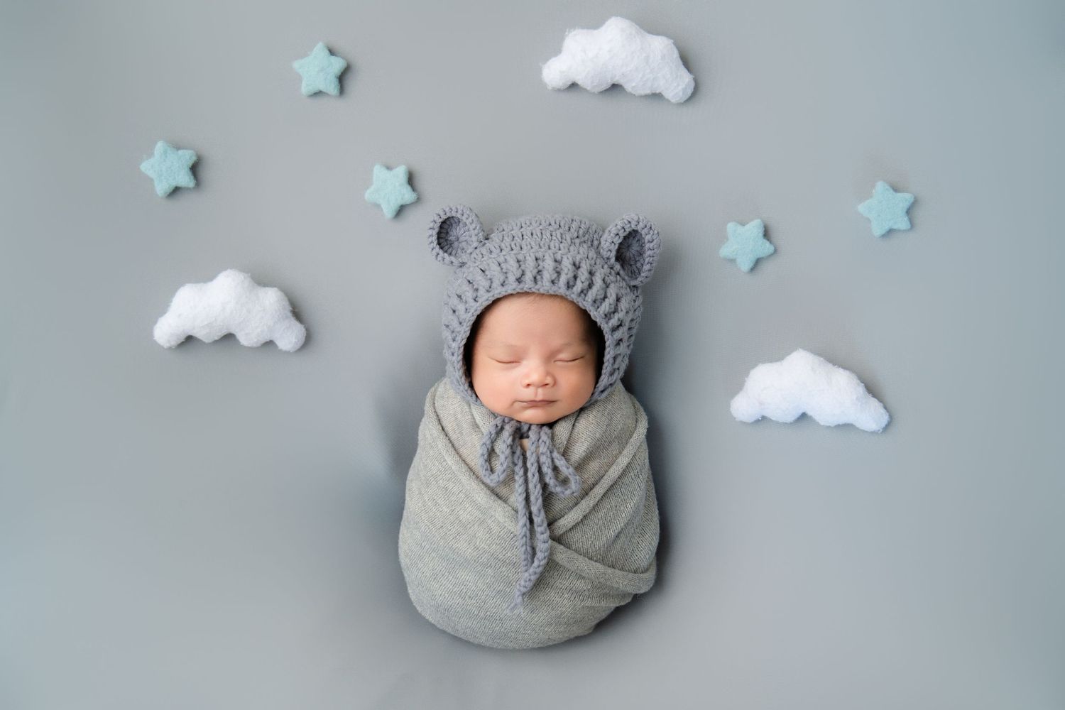 Newborn in grey bear hat sleeping among paper clouds and stars on grey backdrop.