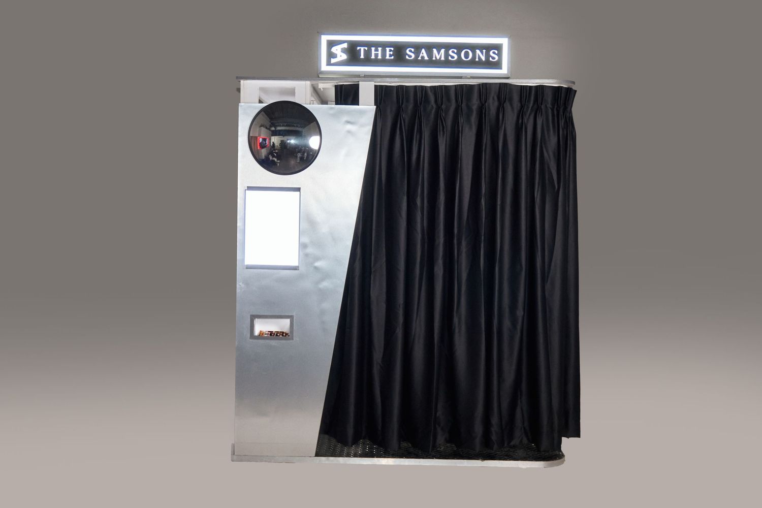 The Samsons branded photo booth with black curtain and camera lens on gray background.