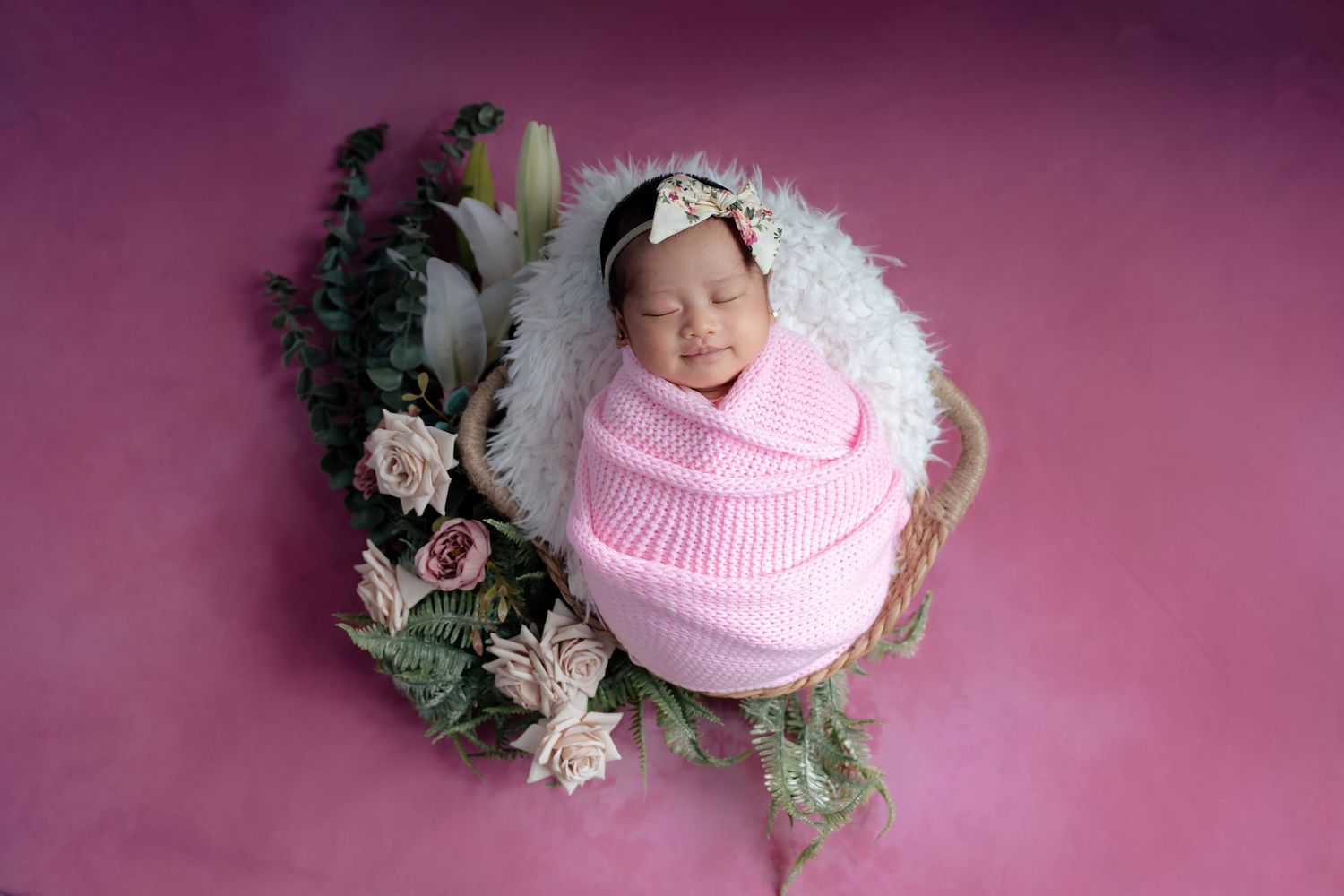Newborn wrapped in pink blanket sleeping in floral basket against pink background during professional photo session.