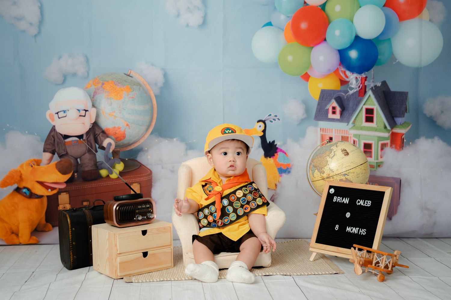 A creative travel-themed photography setup with colorful balloons, vintage suitcases, and a model house.