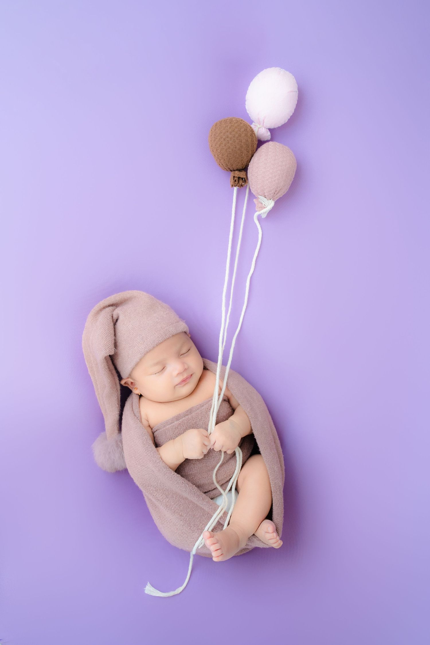Sleeping newborn in beige outfit holding balloon props against purple background.