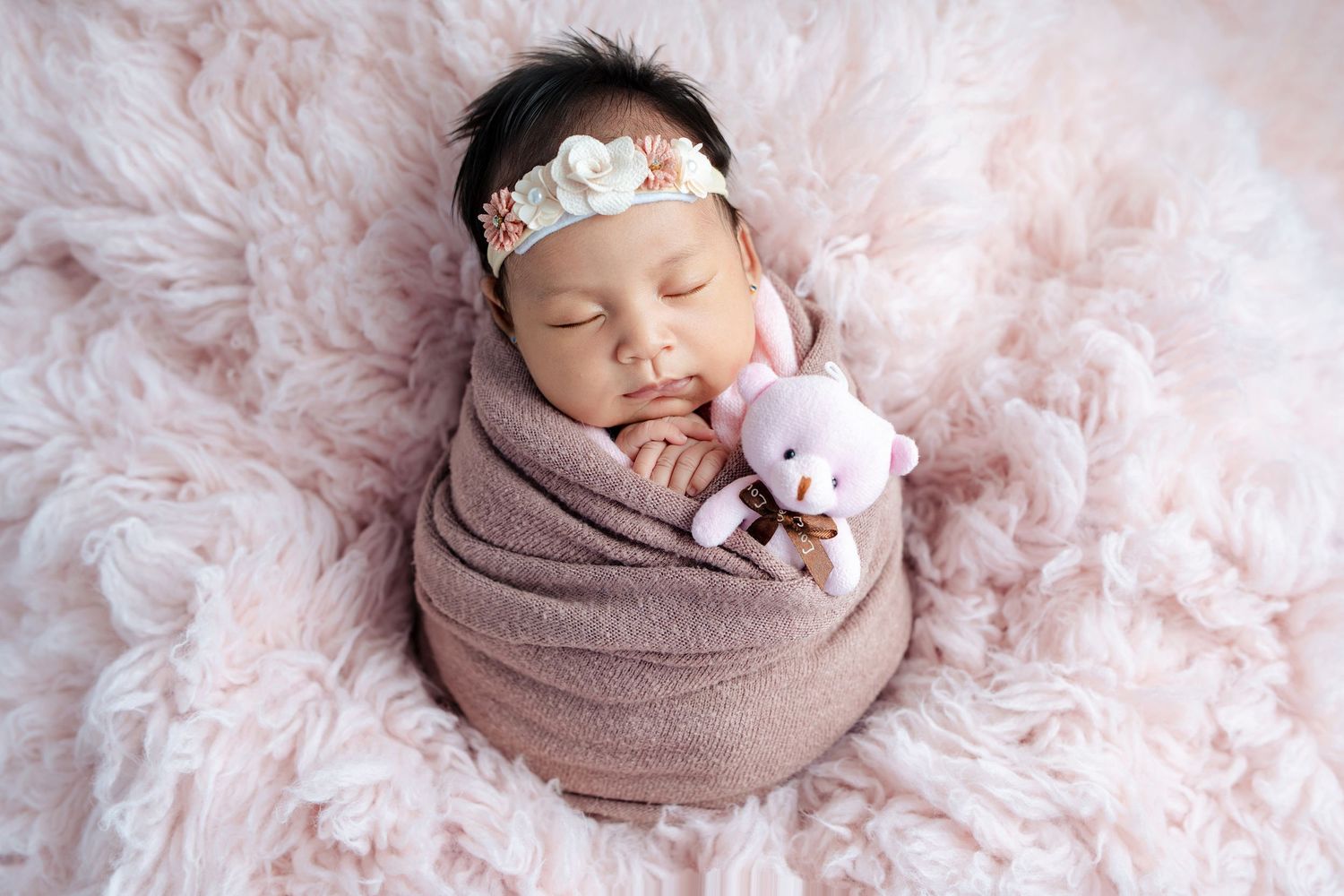 A sleeping newborn wrapped in soft mauve fabric lies on a pink floral backdrop with a delicate tiara and plush toy.