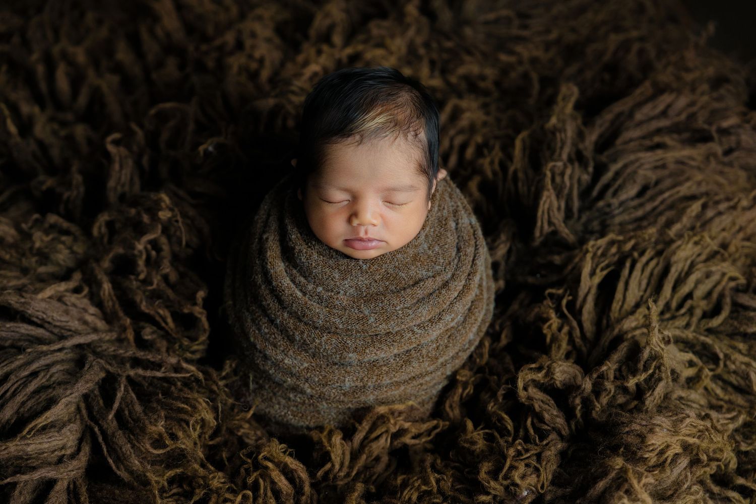 Swaddled newborn sleeping on textured brown fabric during studio photography session.