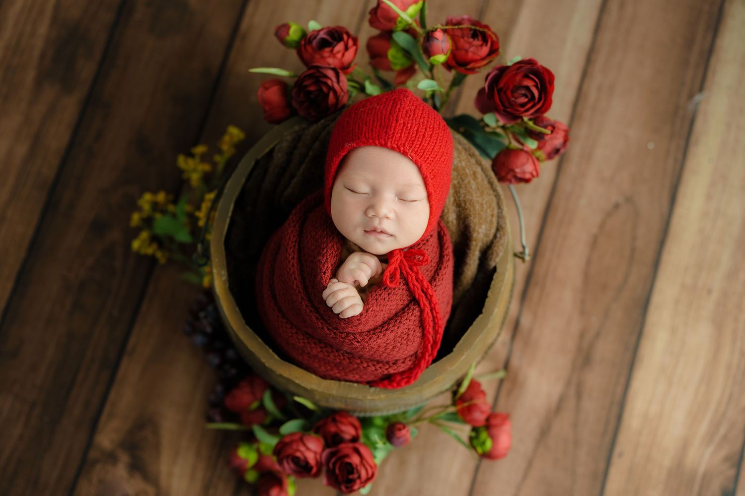 Newborn in red wrap sleeping in wooden bowl surrounded by red roses and greenery.