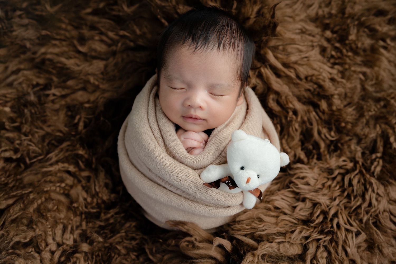 Newborn baby wrapped in beige swaddle sleeping peacefully on brown fur blanket with small white teddy bear.