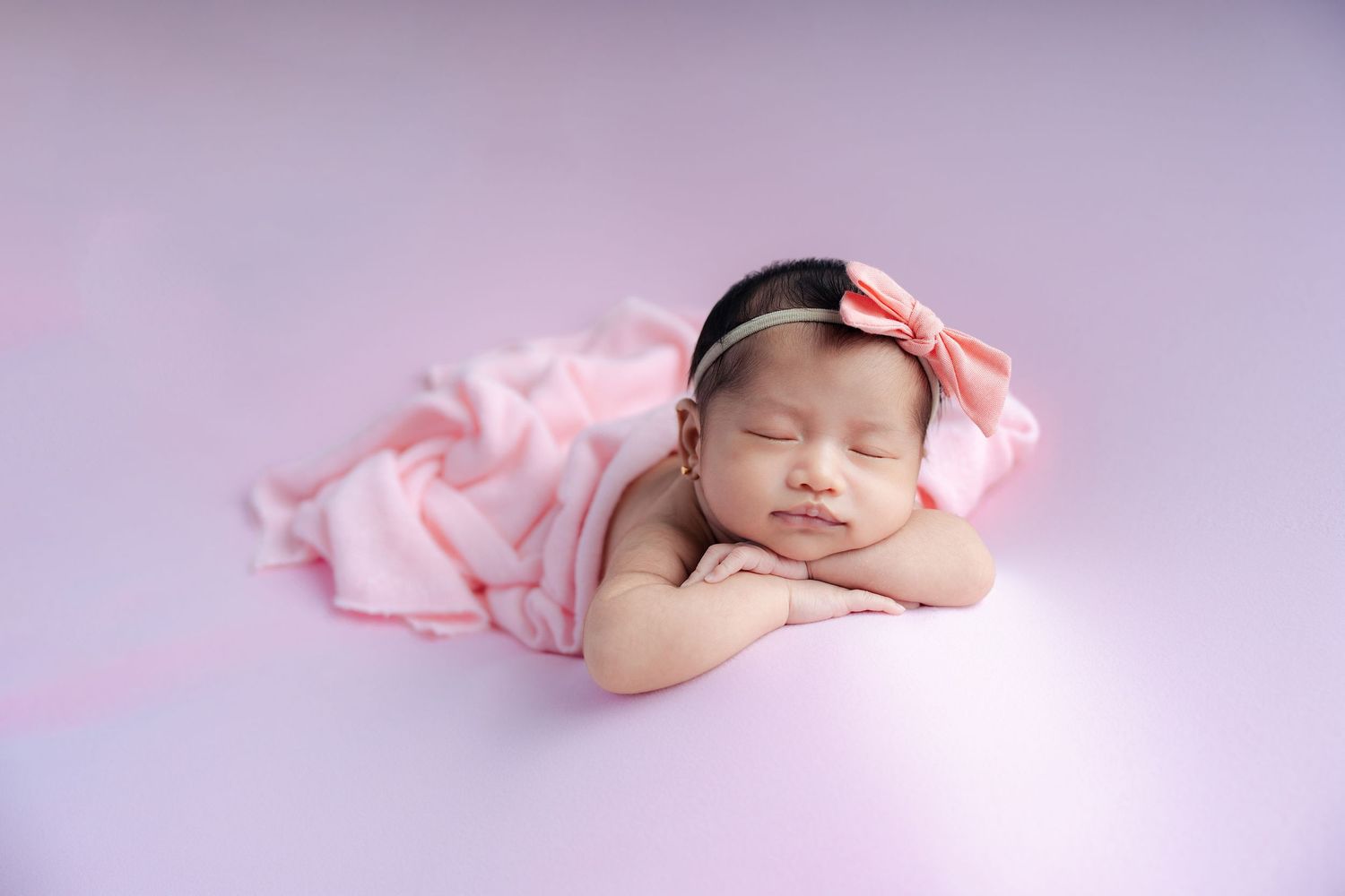 Newborn baby sleeping peacefully in pink swaddle blanket on soft backdrop during professional photo session.