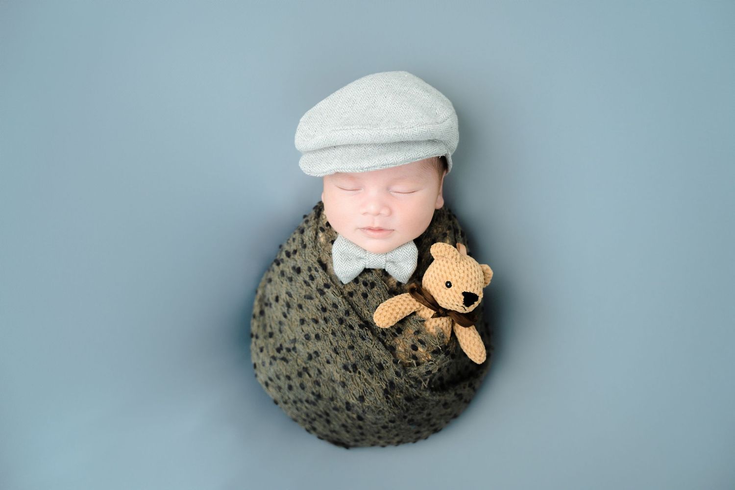 Newborn wearing white cap photographed with teddy bear against light blue background.
