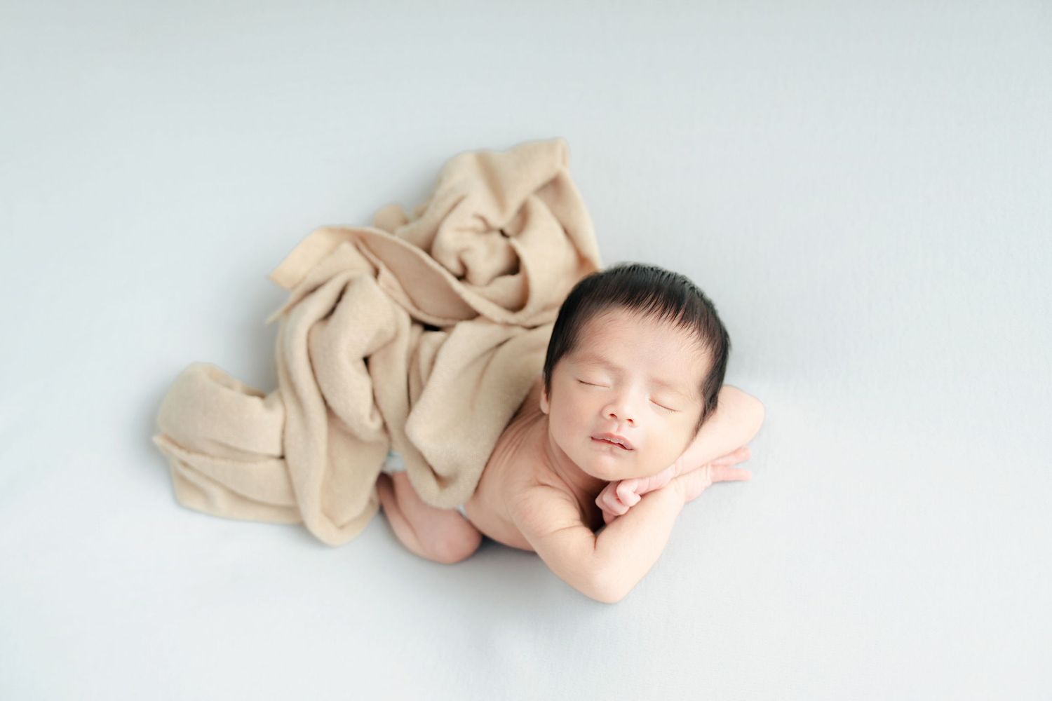 Peaceful sleeping newborn baby wrapped in beige fabric on light background during professional photo session.