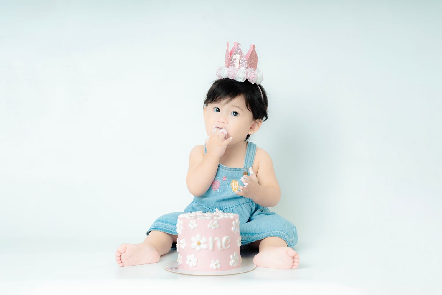 Baby wearing bunny ears sits next to pink birthday cake on white background during cake smash photo session.