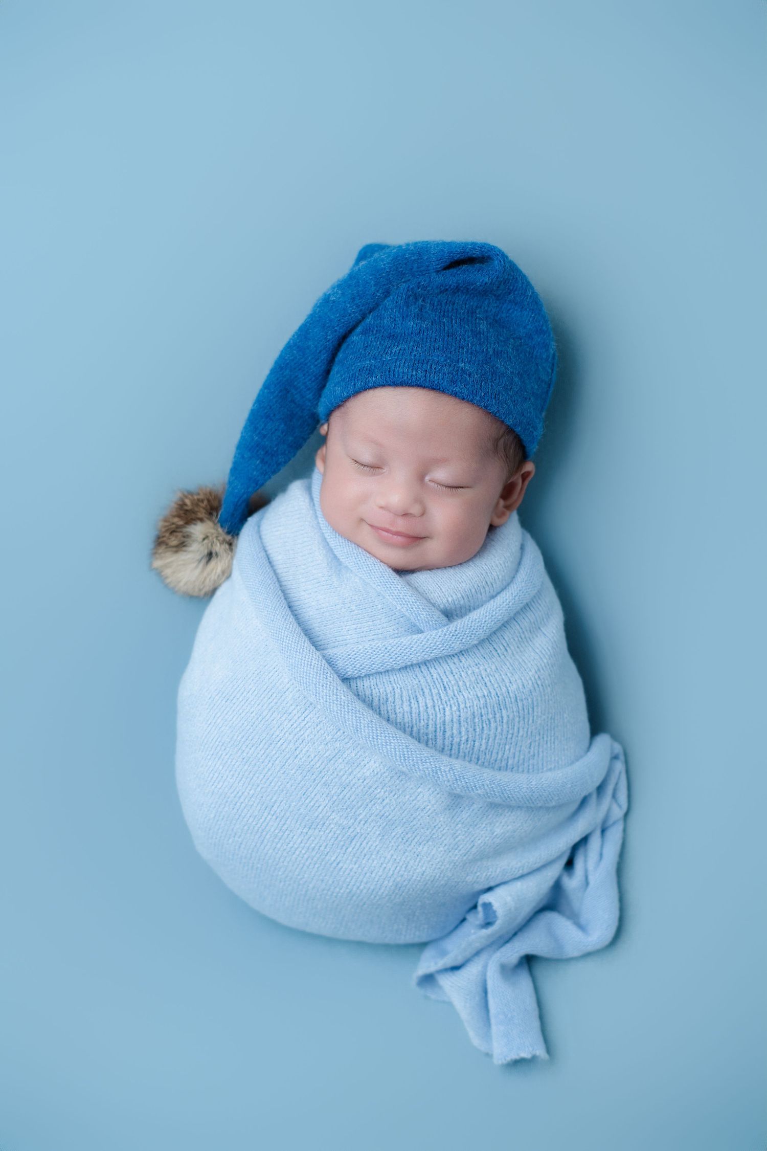 Sleeping newborn in blue hat and wrap against light blue background.