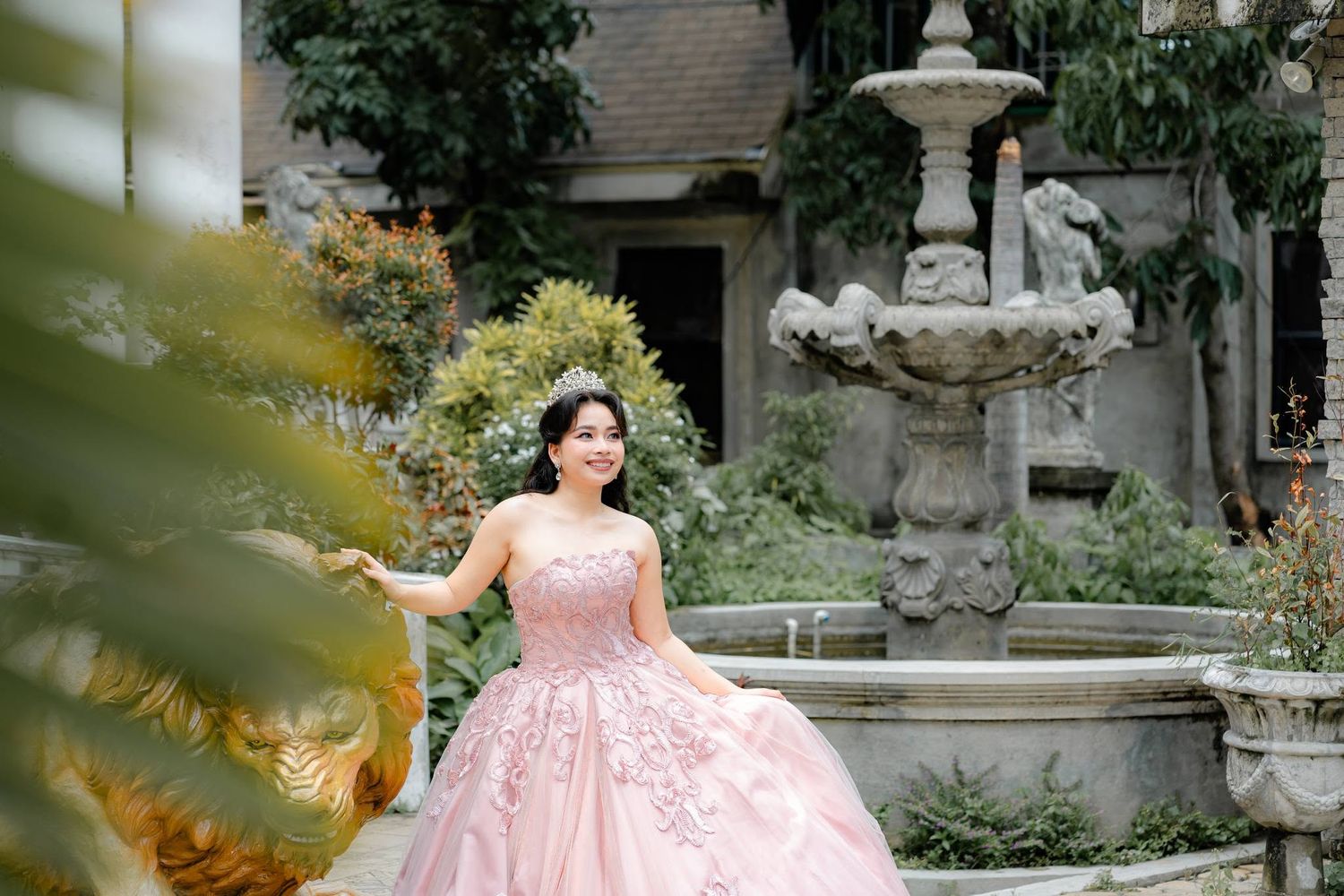 Young woman in a pink ball gown poses gracefully by a stone fountain in a lush garden setting.
