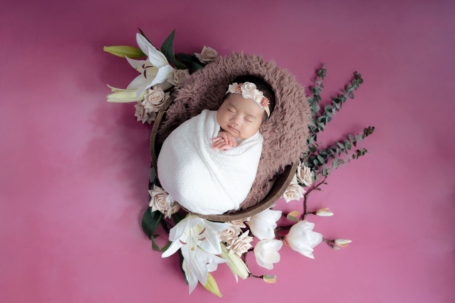 Overhead view of newborn baby wrapped in white swaddle lying in round nest with pink flowers and lavender on pink background.