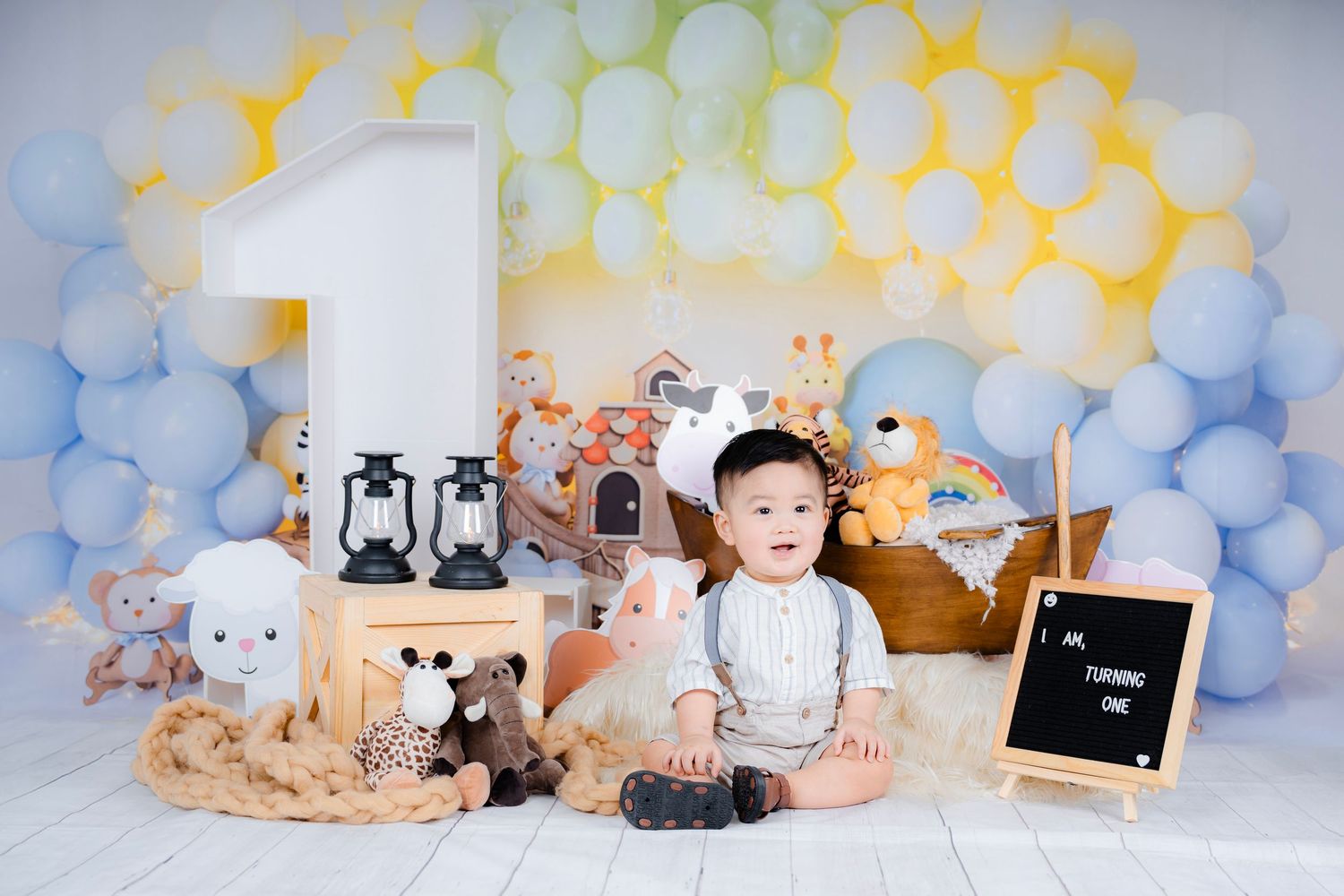 First birthday milestone photo with soft blue and yellow balloon arch and stuffed animals.