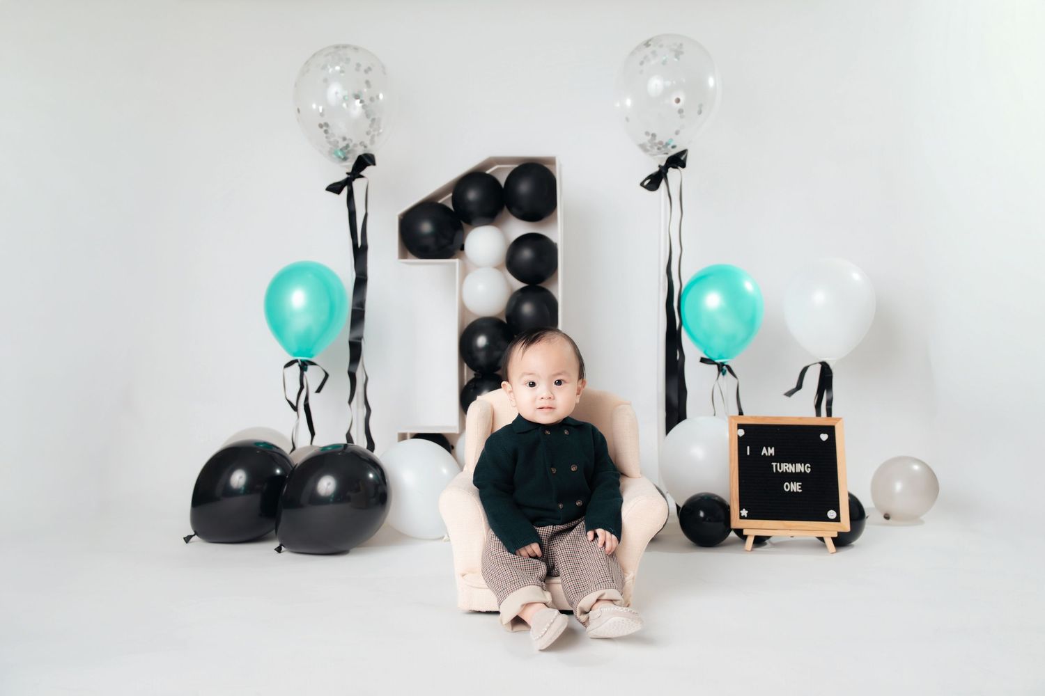 First birthday celebration setup with black and teal balloons, number one balloon, and decorative elements on white background.