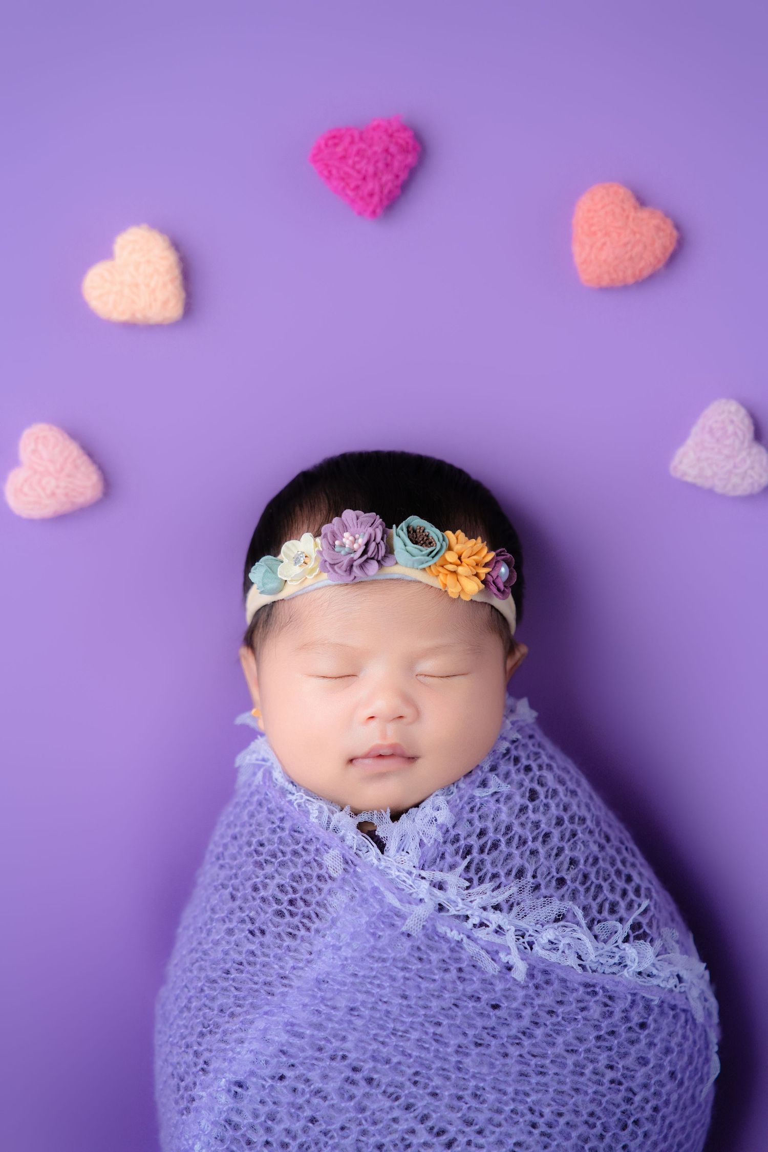 Swaddled baby in purple blanket with floral headband and felt hearts on purple backdrop.