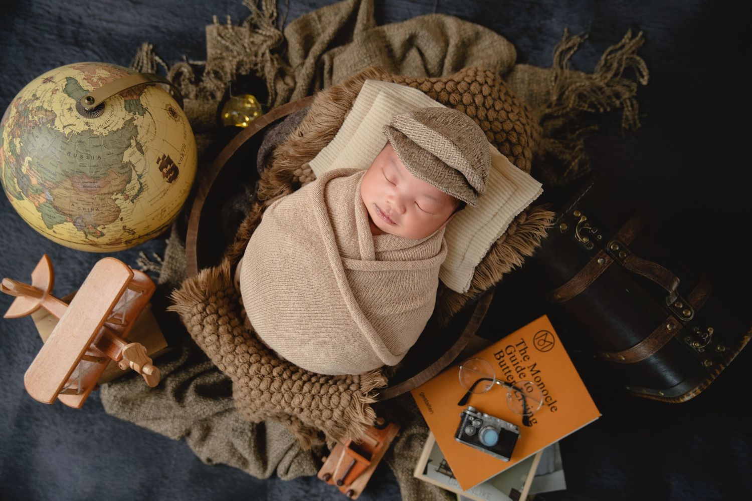 Vintage travel themed newborn photo with globe, wooden airplane, and beige wrapping on dark background.