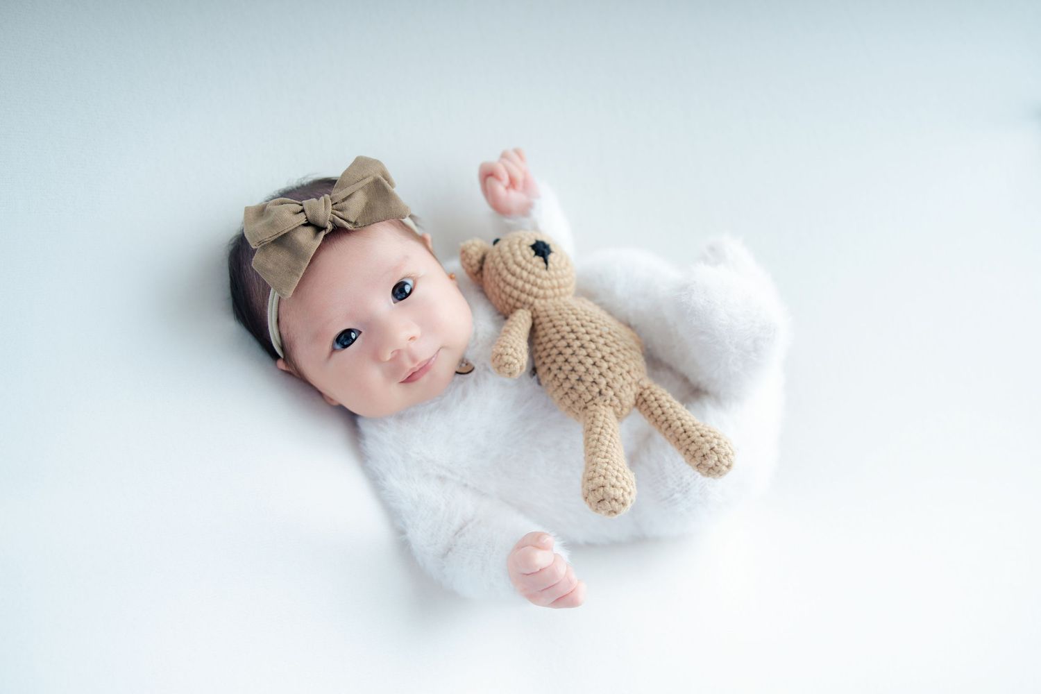 A small infant lying on soft white bedding with a knitted brown teddy bear companion.