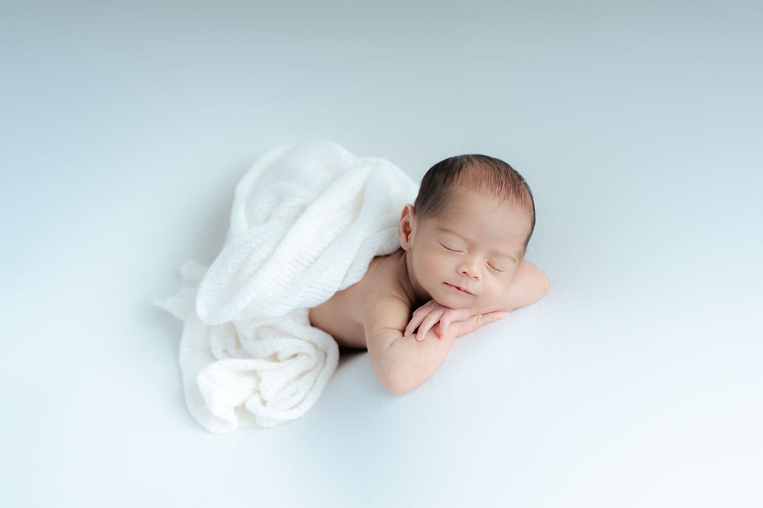 A sleeping newborn baby wrapped in soft white fabric lies peacefully on a light background.