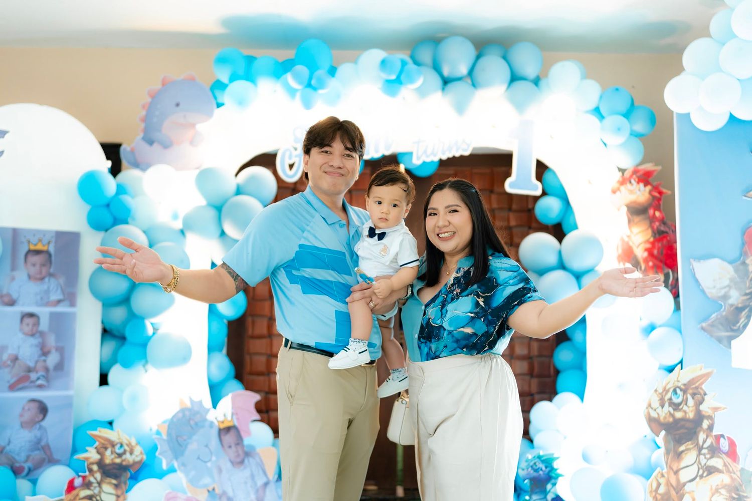 Family celebrating baby's first birthday with blue balloon arch backdrop at a festive party venue.