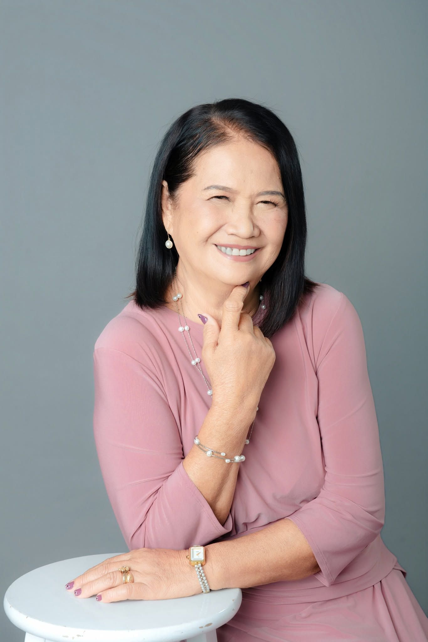 Smiling middle-aged Asian woman in pink dress with jewelry, posing confidently against gray background.