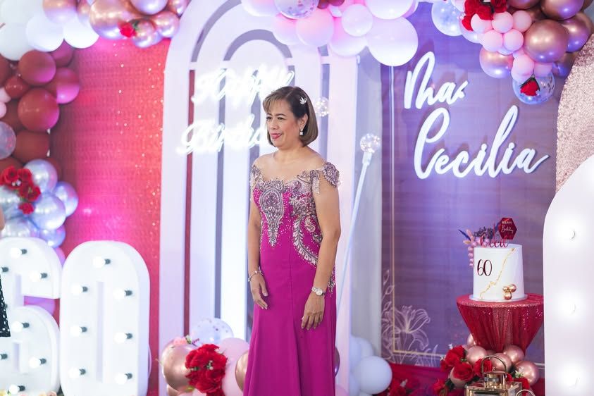Woman in elegant pink gown poses at Thas Cecilia 60th birthday party with balloon arch and cake.