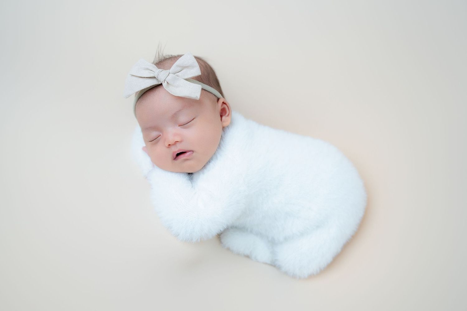 Sleeping newborn in white wrap with bow headband on cream background.