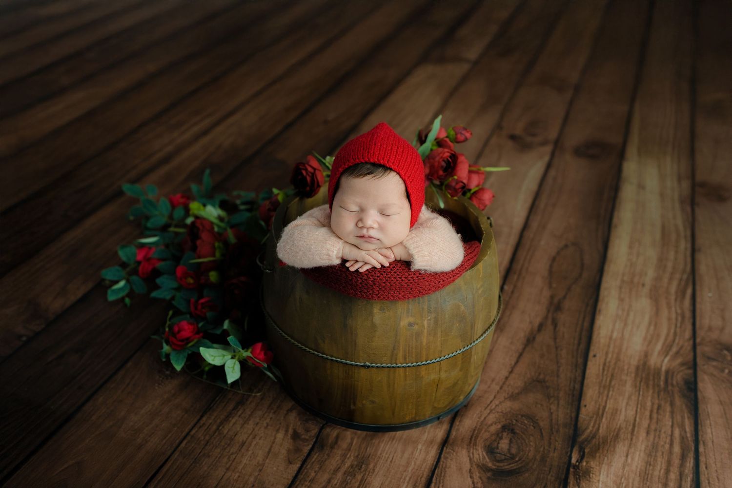 Sleeping newborn in red cap posed in rustic wooden barrel with holiday greenery.