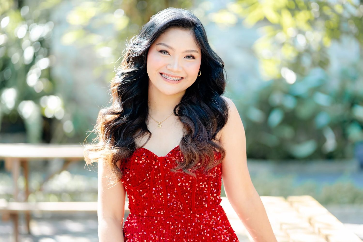 Smiling young woman in a red sequin dress poses outdoors among lush greenery in a professional portrait.