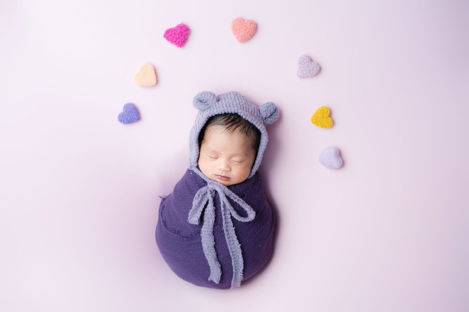 Sleeping newborn in navy wrap with bear bonnet surrounded by colorful heart decorations.
