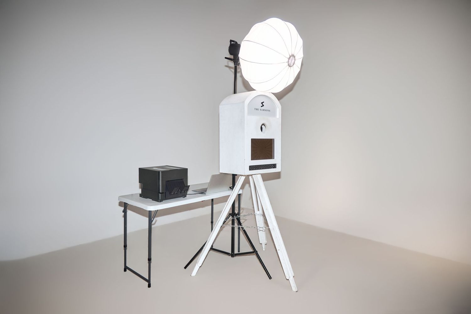 White photo booth on tripod with large round flash and black printer on folding table against gray wall.