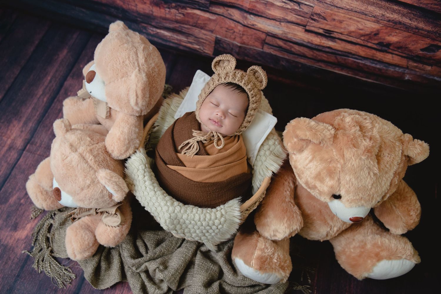 Sleeping baby in bear hat nestled among large teddy bears on rustic wooden backdrop.