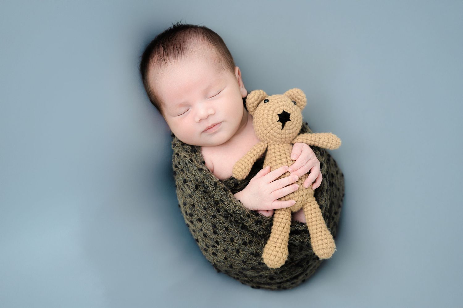 Sleeping baby cuddling small brown teddy bear on blue backdrop.